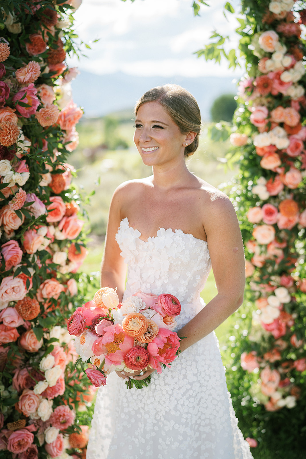 Bride in white floral gown holding bouquet outdoors.