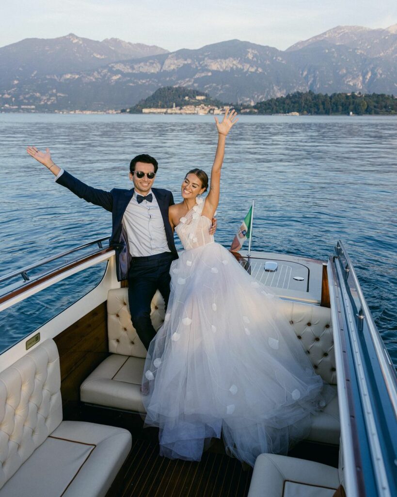 Bride and groom celebrate on boat, mountains behind.