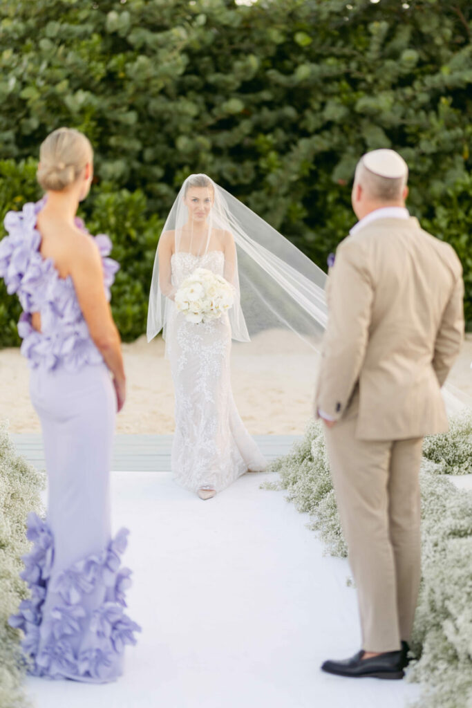 Bride in couture gown walks outdoor aisle.