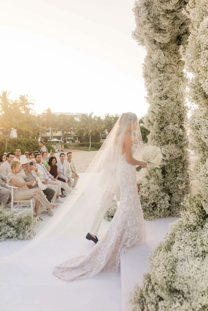 Bride in couture gown walks floral outdoor aisle.