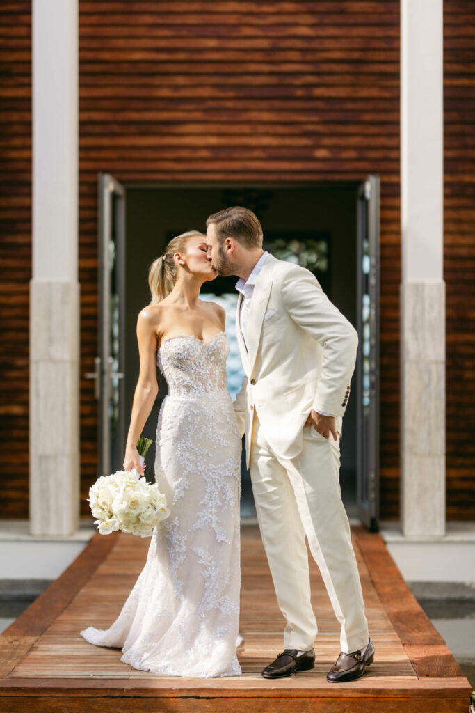 Bride kisses groom on walkway, holding bouquet.