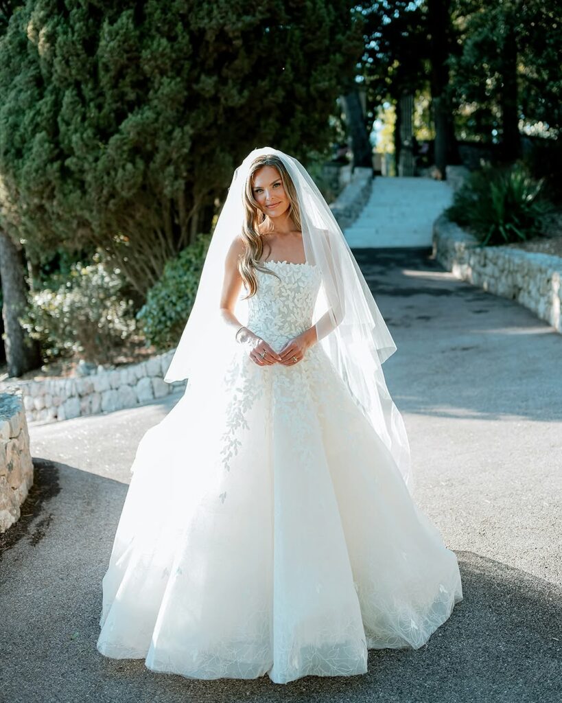 Bride in white gown smiles on sunlit path.