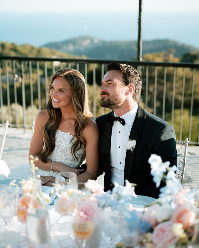 Smiling newlyweds at outdoor table, scenic backdrop.