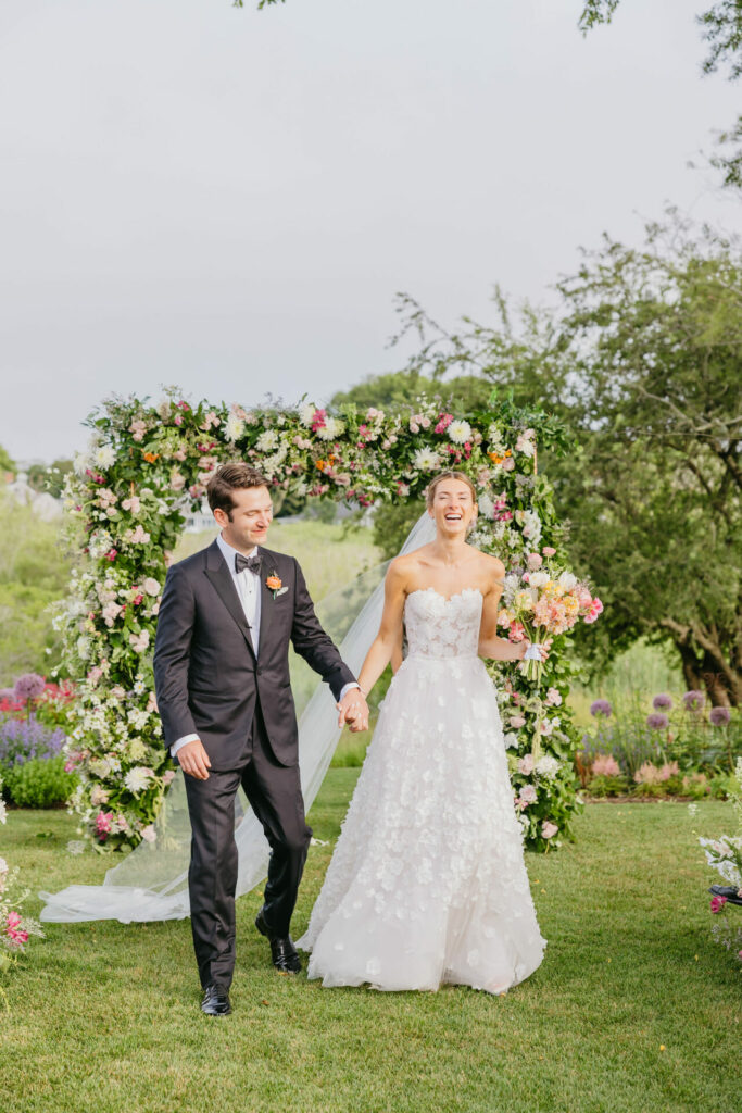 Bride and groom walk outdoors by floral arch.