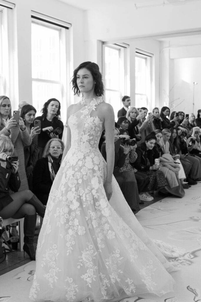 Model in floral lace gown walks runway.