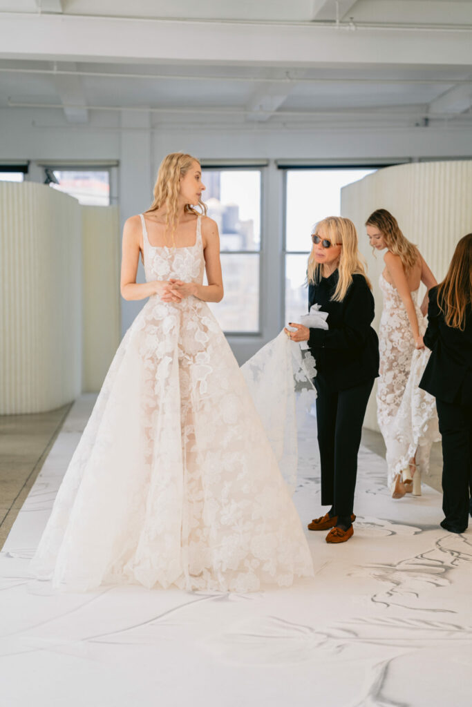 Model in white floral wedding dress, others adjusting.