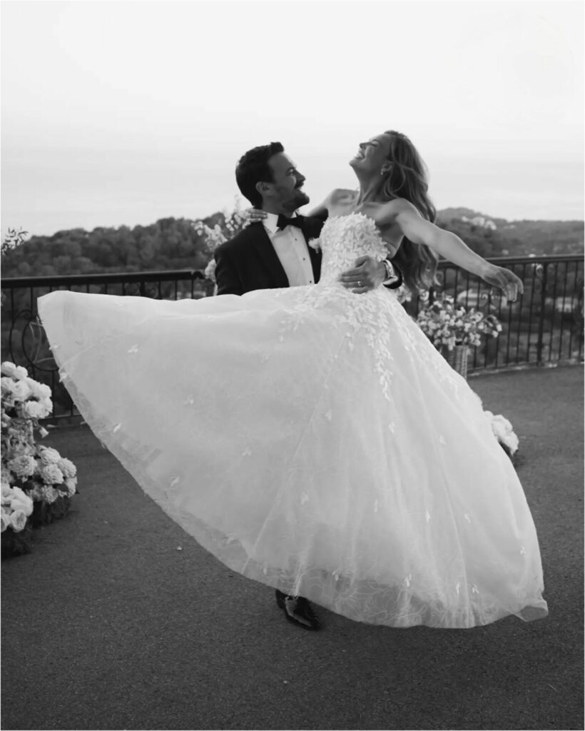 Groom lifts smiling bride outdoors amid flowers.