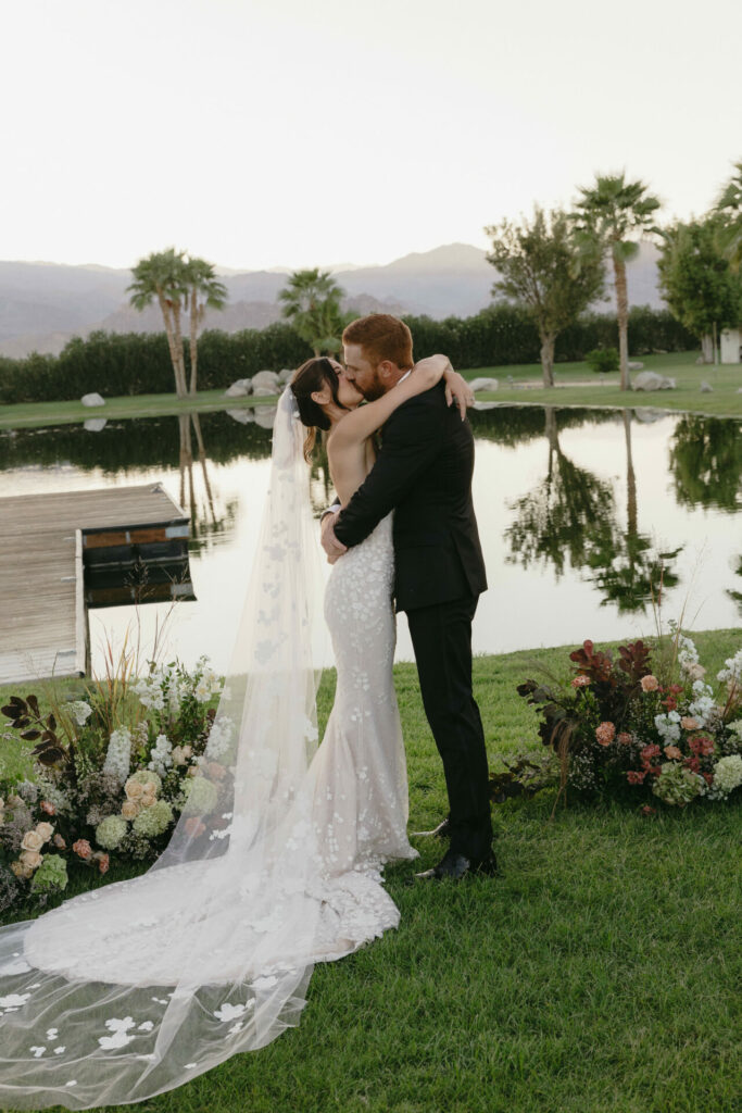 Bride and groom kiss by lake, floral veil.