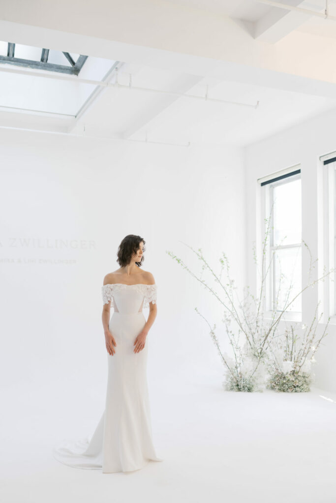 Bride in white dress stands by large windows.