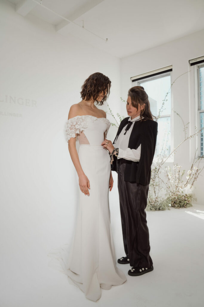 Bride in white dress, suit-wearing woman assists.