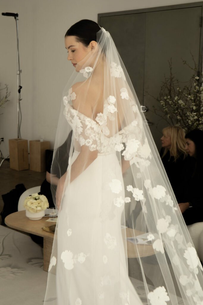 Bride in white gown, floral veil indoors.