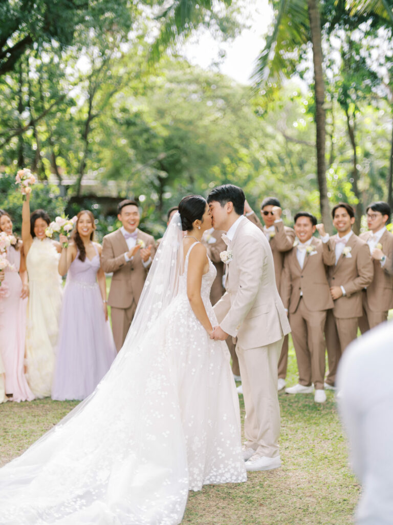Bride and groom kiss, wedding party cheers outdoors.