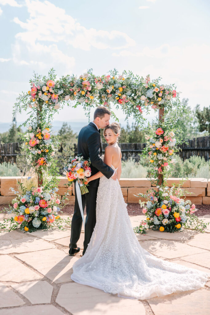 Bride and groom embrace under floral arch outdoors.