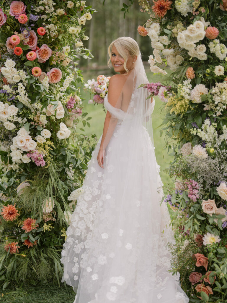 Bride in floral dress stands by flower arches.