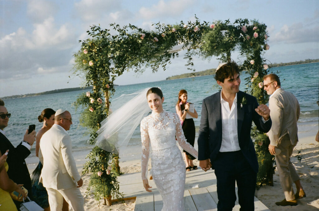 Bride and groom walk under floral arch, beach.