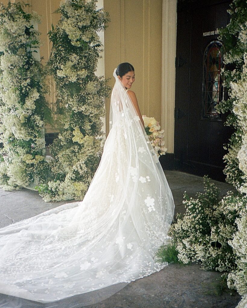 Bride in floral gown, smiling amid white flowers.