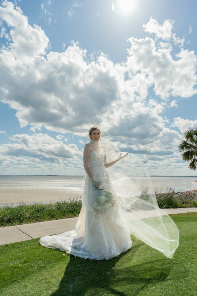 Bride in white holds bouquet by beach.
