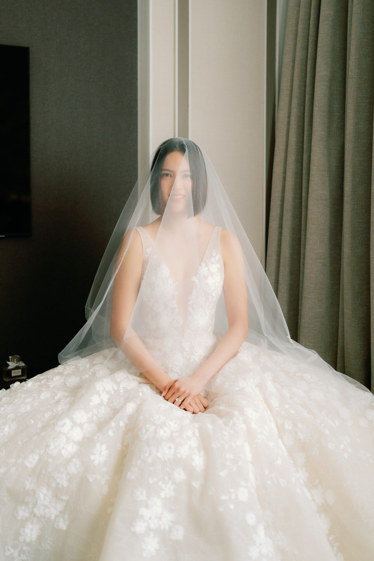 Bride in lace gown and veil smiles, seated indoors.