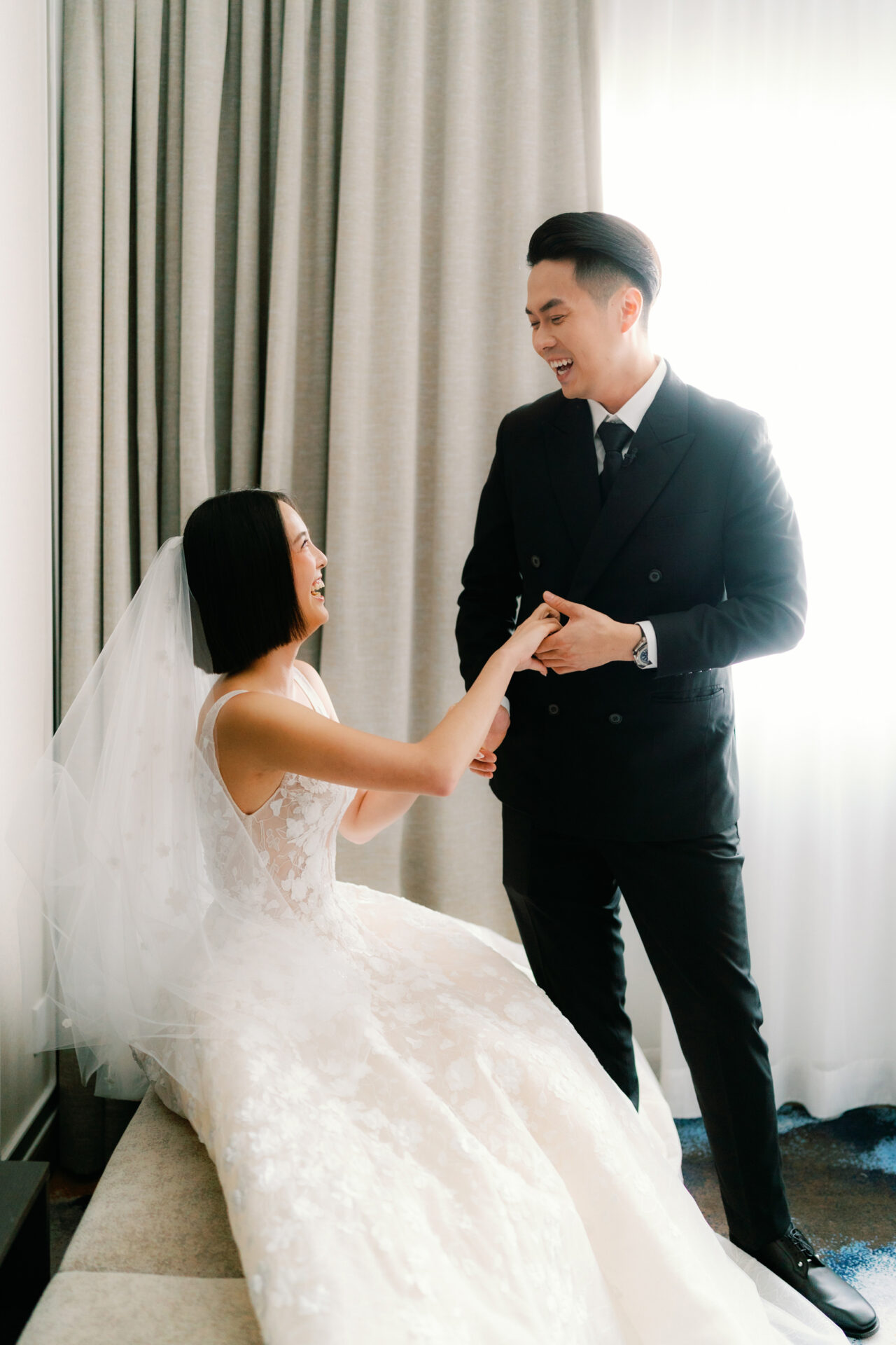Bride smiles on bench, holding groom’s hand.