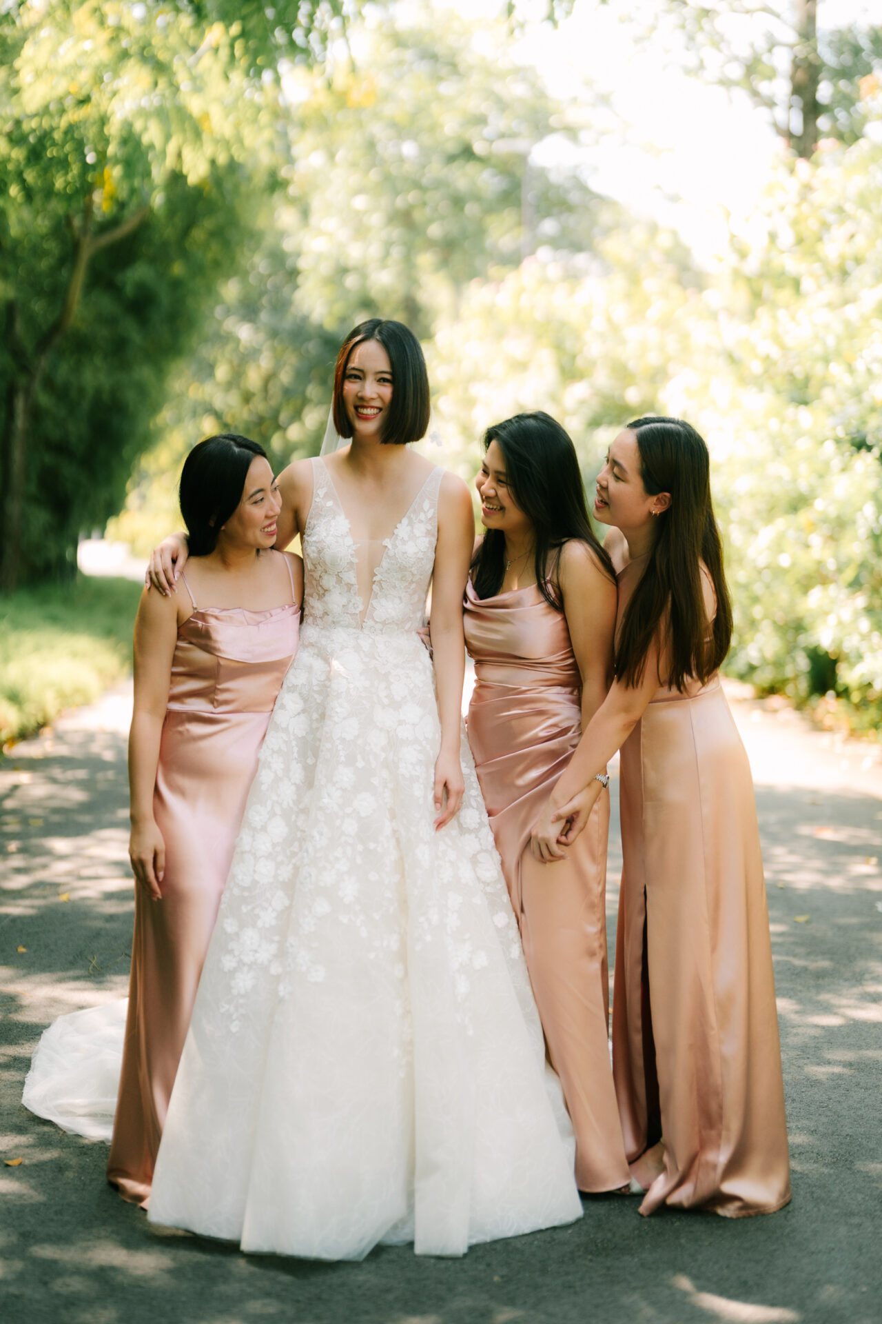 Bride laughs outdoors with three bridesmaids in sunlight.