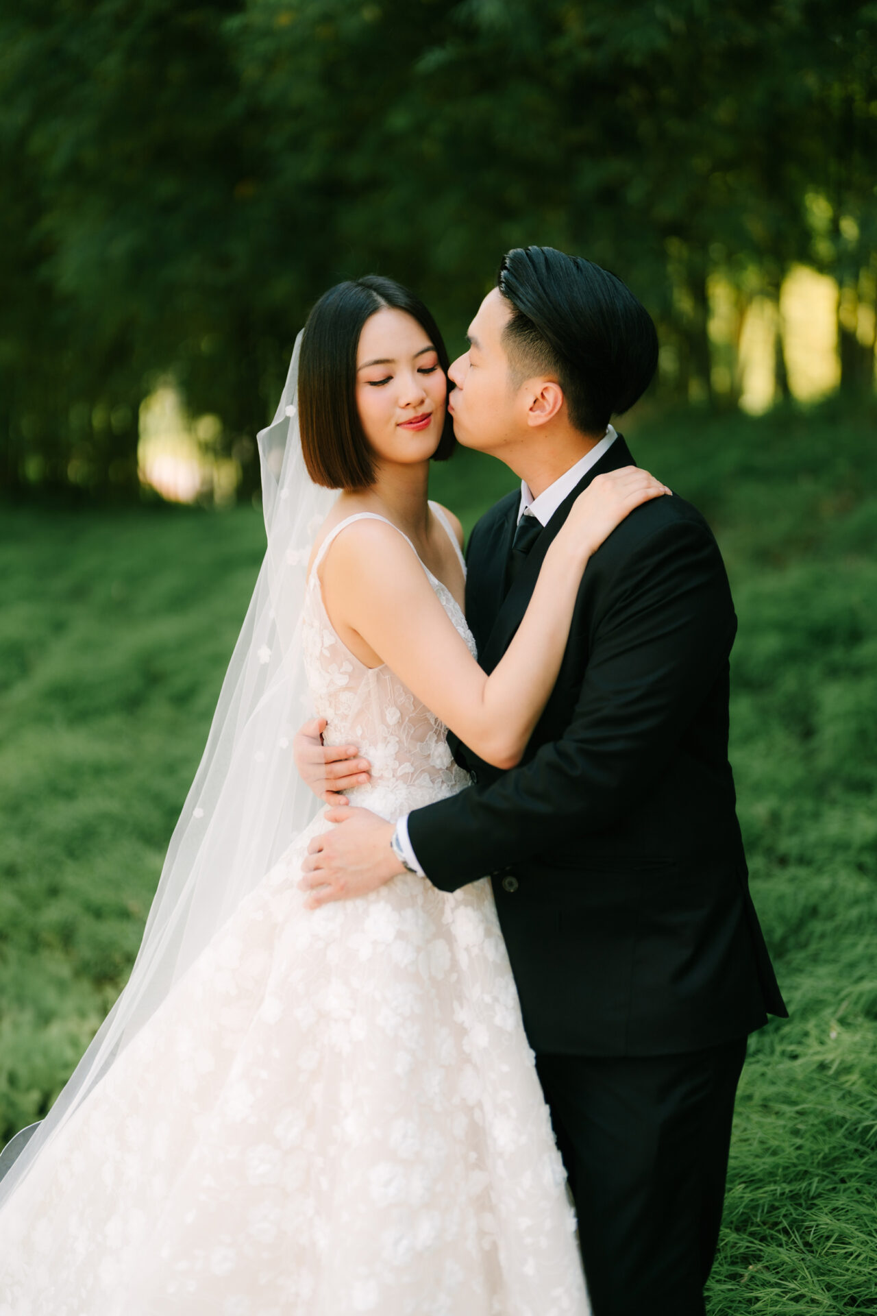 Bride smiles as groom kisses her temple outdoors.