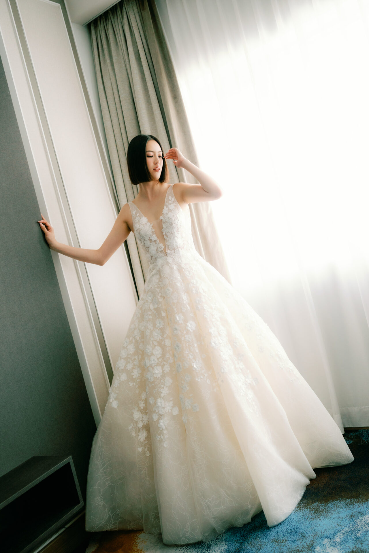 Woman in white floral gown poses by window.