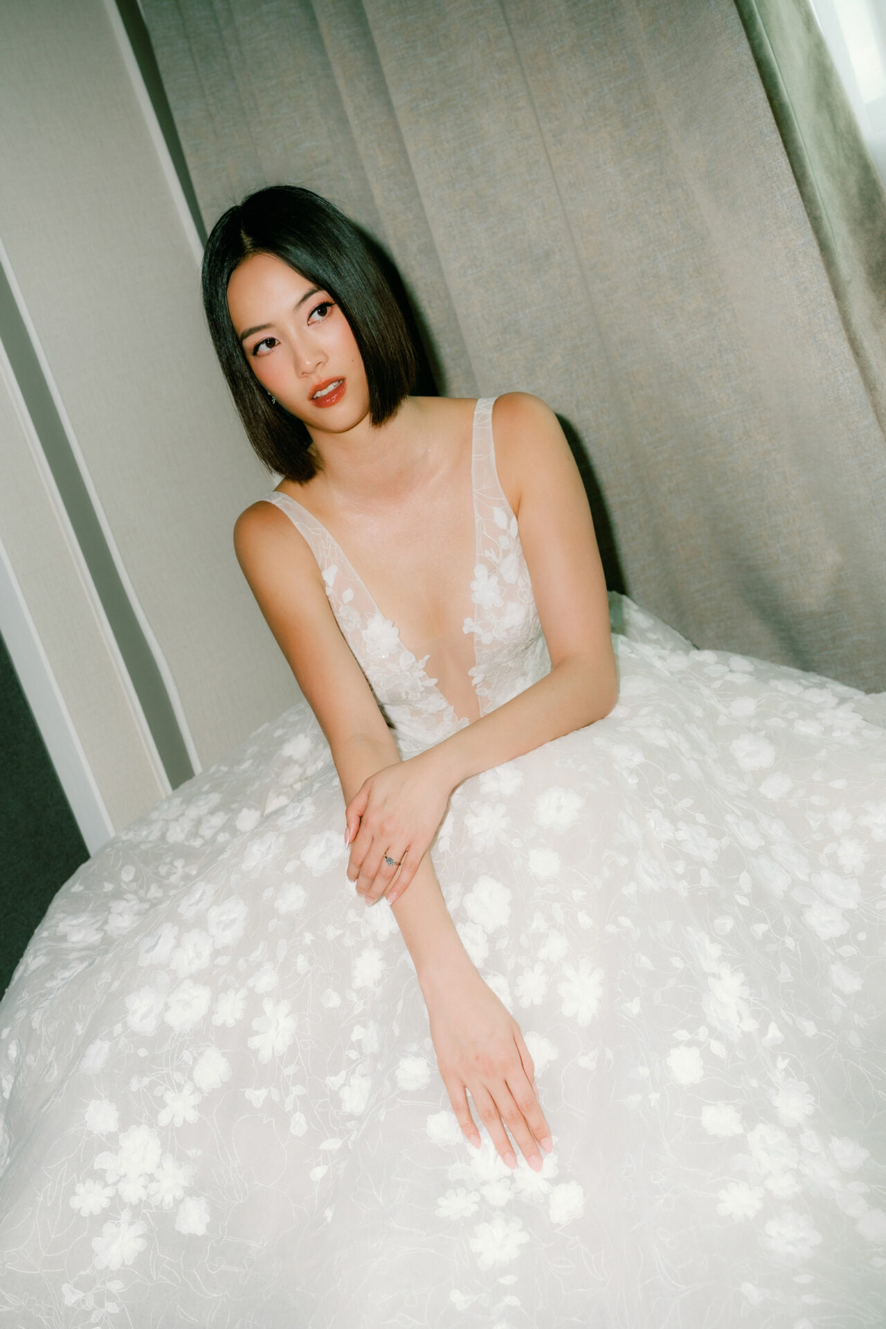 Woman in lace wedding dress sits calmly indoors.