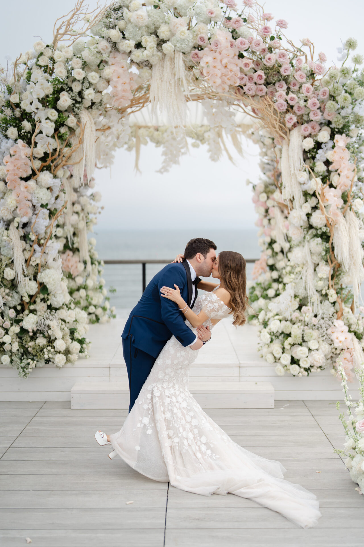 Groom dips and kisses bride under floral arch.