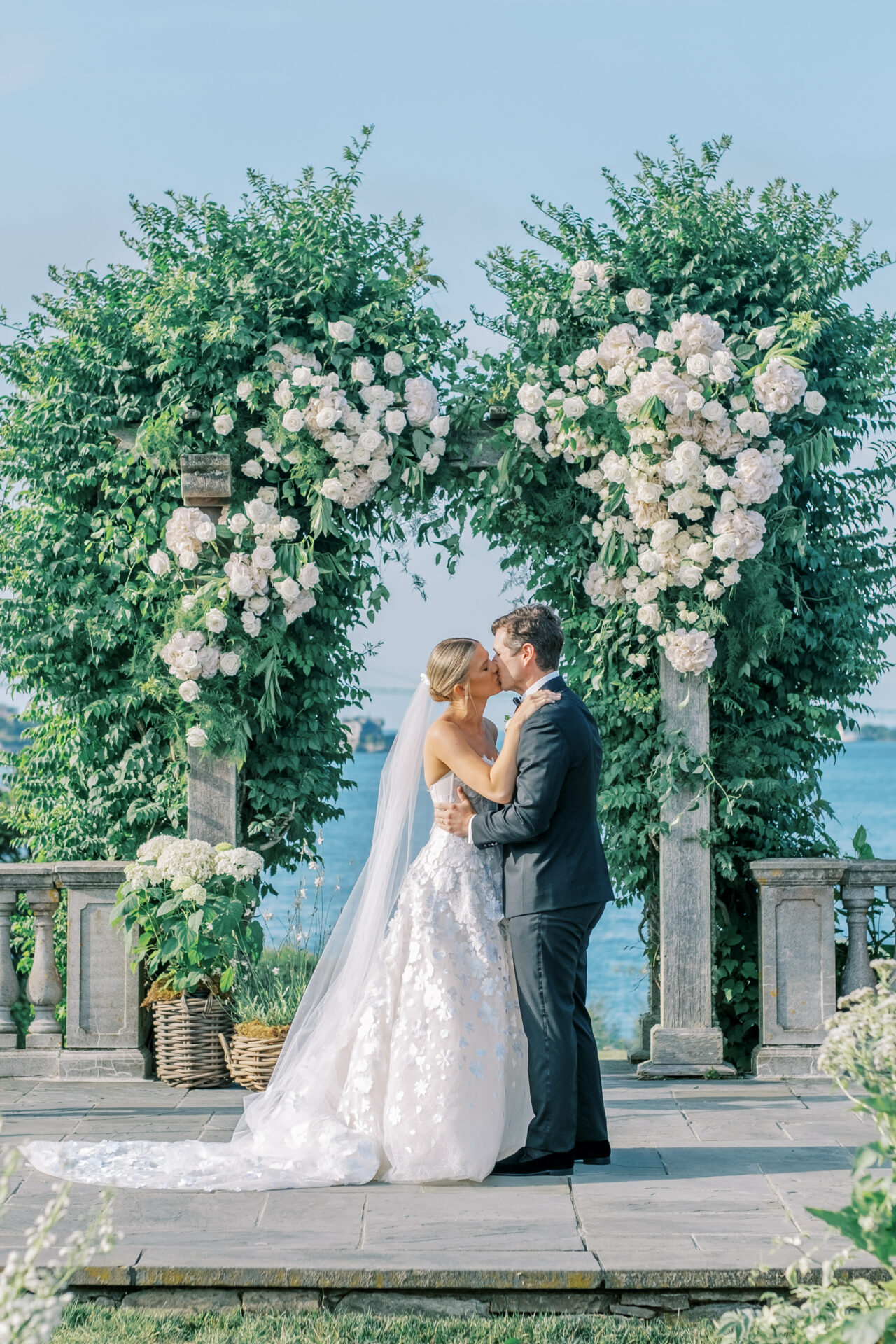 Bride and groom smile under floral arch outdoors.