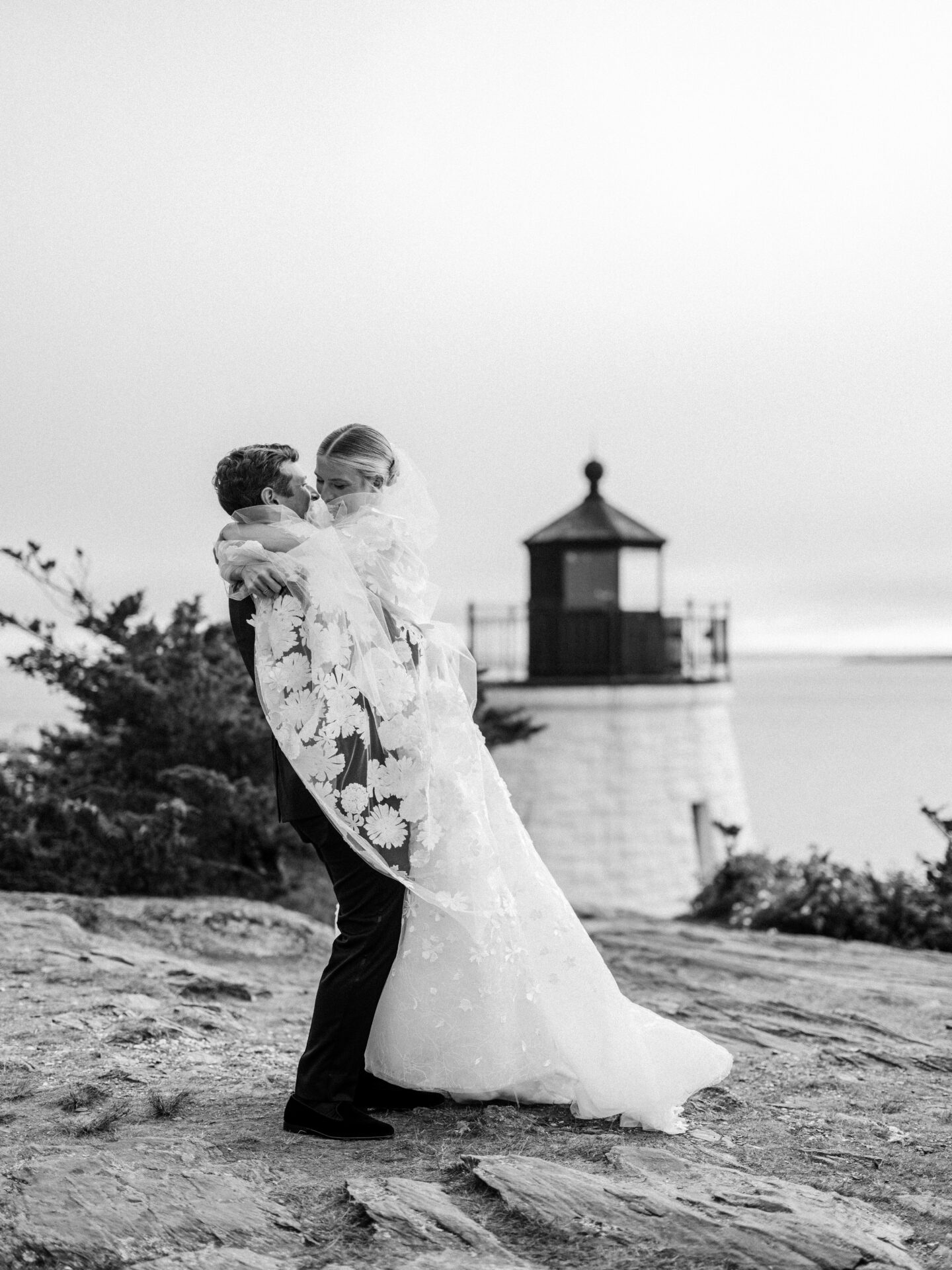Groom lifts bride by lighthouse, black and white.