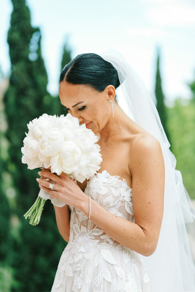 Bride in lace gown with peony bouquet, smiling outdoors.