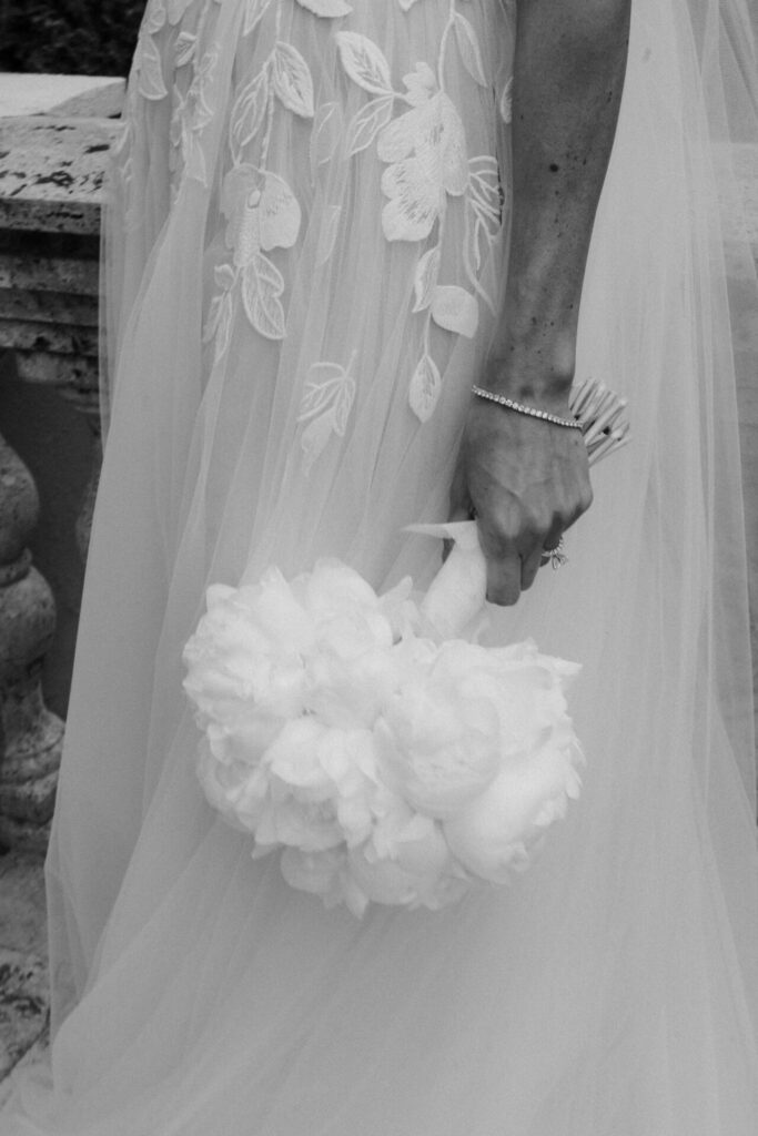 Katya Ruiz in lace Mira Zwillinger gown, holding white flowers.