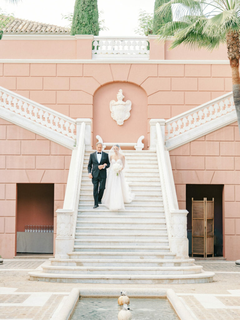 Katya Ruiz in bridal gown descends stairs with partner.