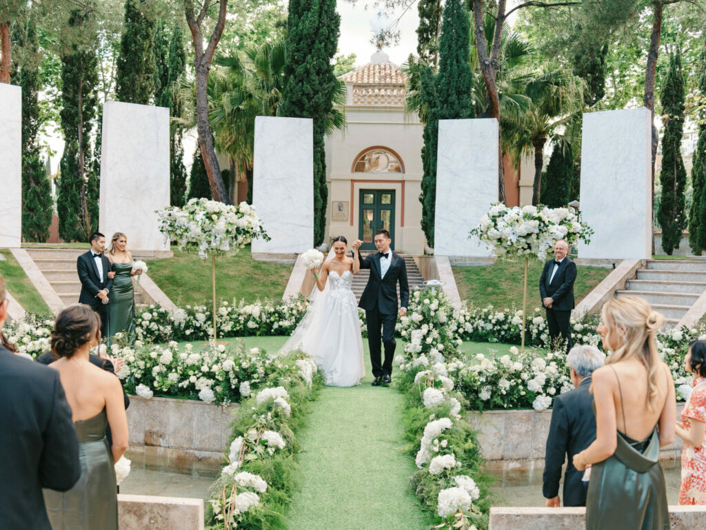 Bride and groom walk outdoor aisle, white flowers everywhere.