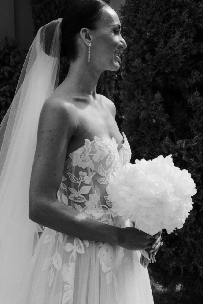 Katya Ruiz smiles in floral gown, holding bouquet outdoors.