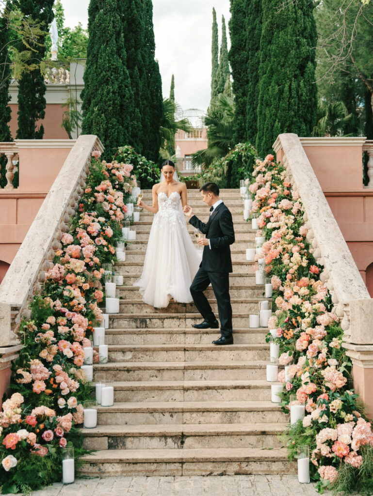 Bride and groom on flower-lined stone stairs, elegant scene.