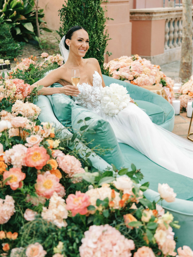 Katya Ruiz in couture gown on green sofa, blooms, champagne.