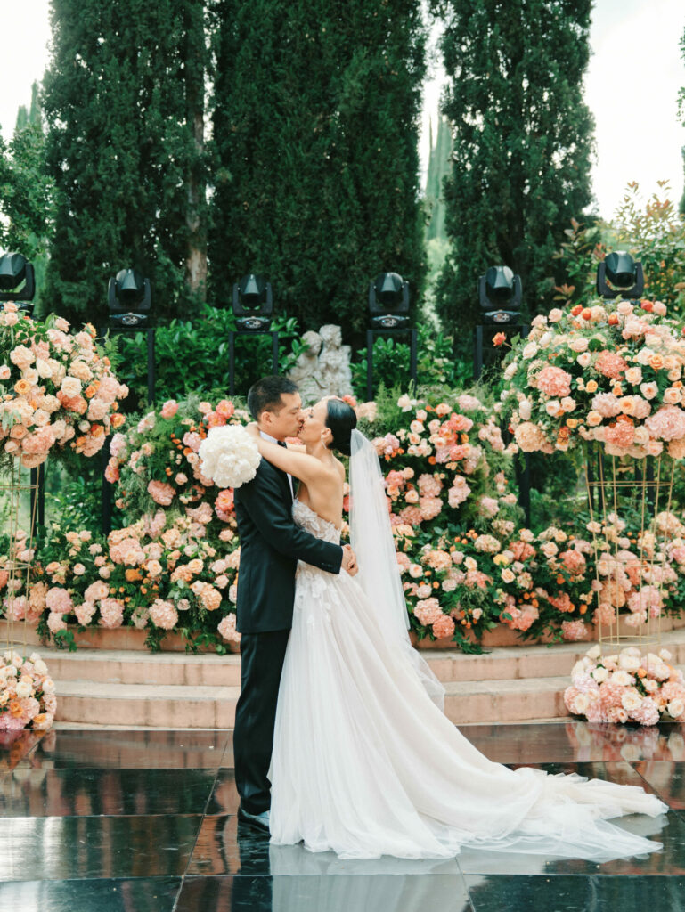 Katya Ruiz kisses groom outdoors, lush blooms, couture gown.