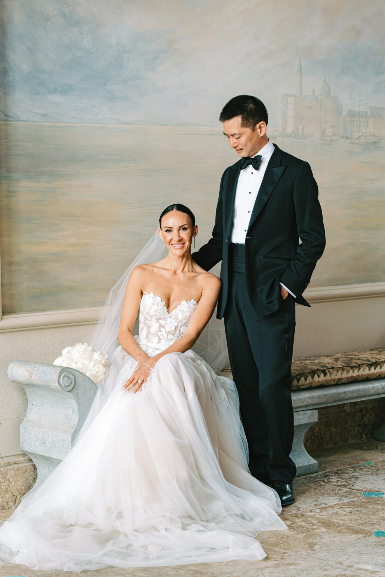 Bride in couture gown sits with bouquet, groom stands.