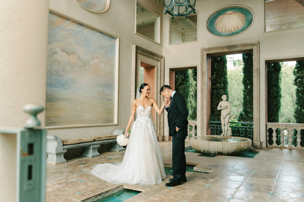 Groom kisses bride’s hand in elegant hallway, smiling.