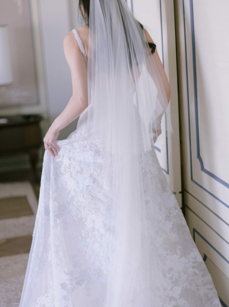Bride in lace gown holds dress, facing away.