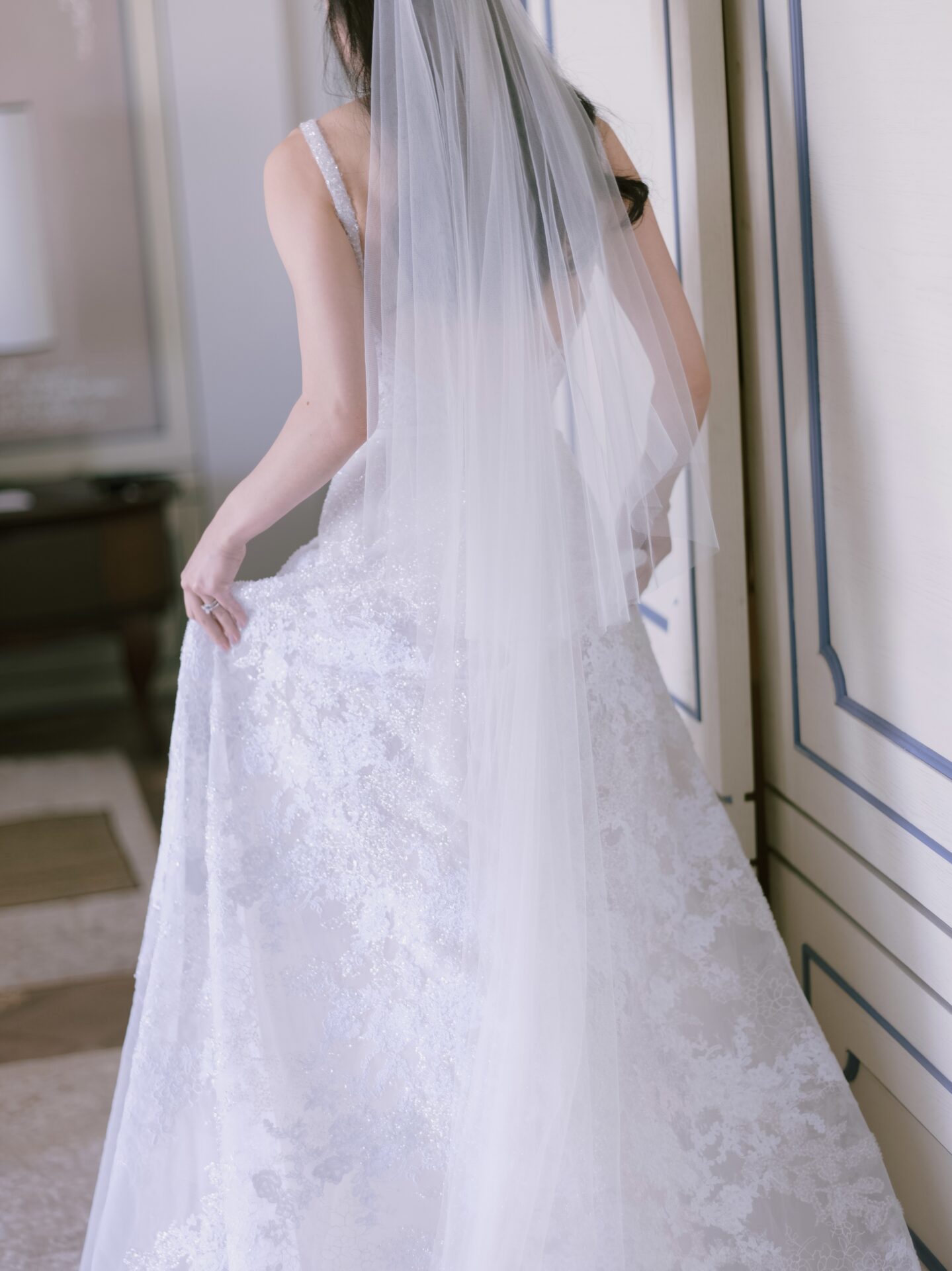 Bride in lace gown holds dress, facing away.