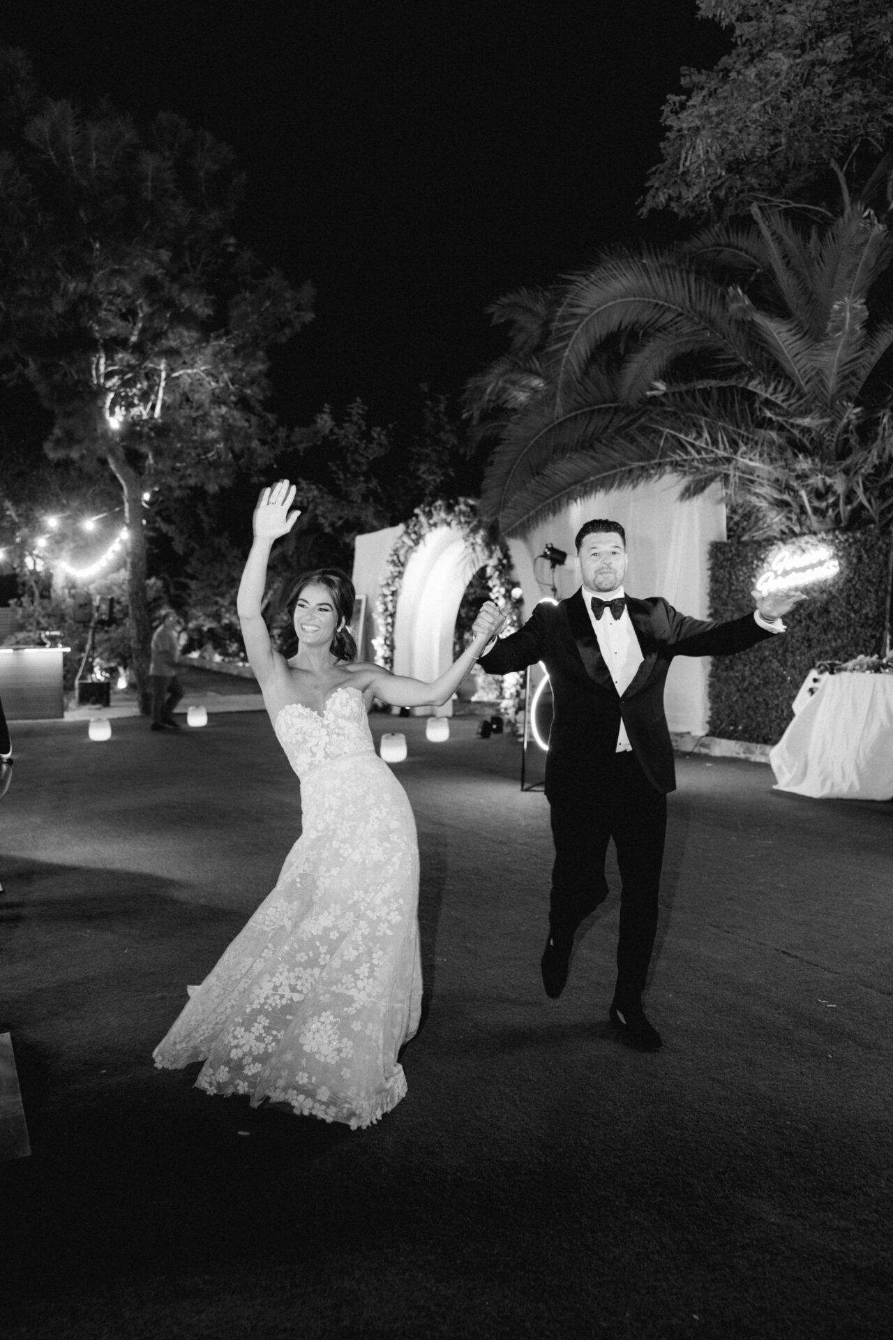 Newlyweds run and wave under night string lights.