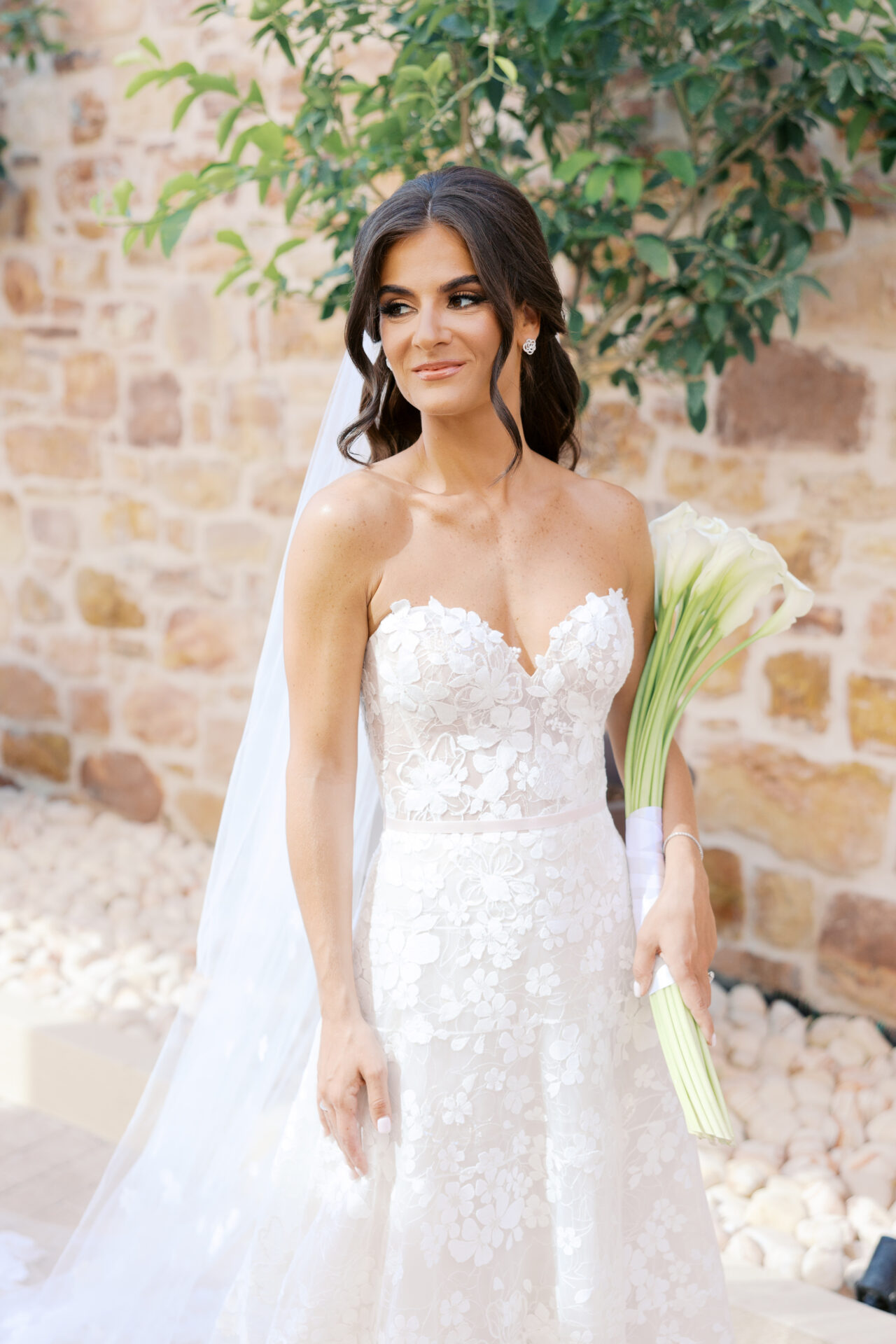 Bride in lace gown with lilies by stone wall.