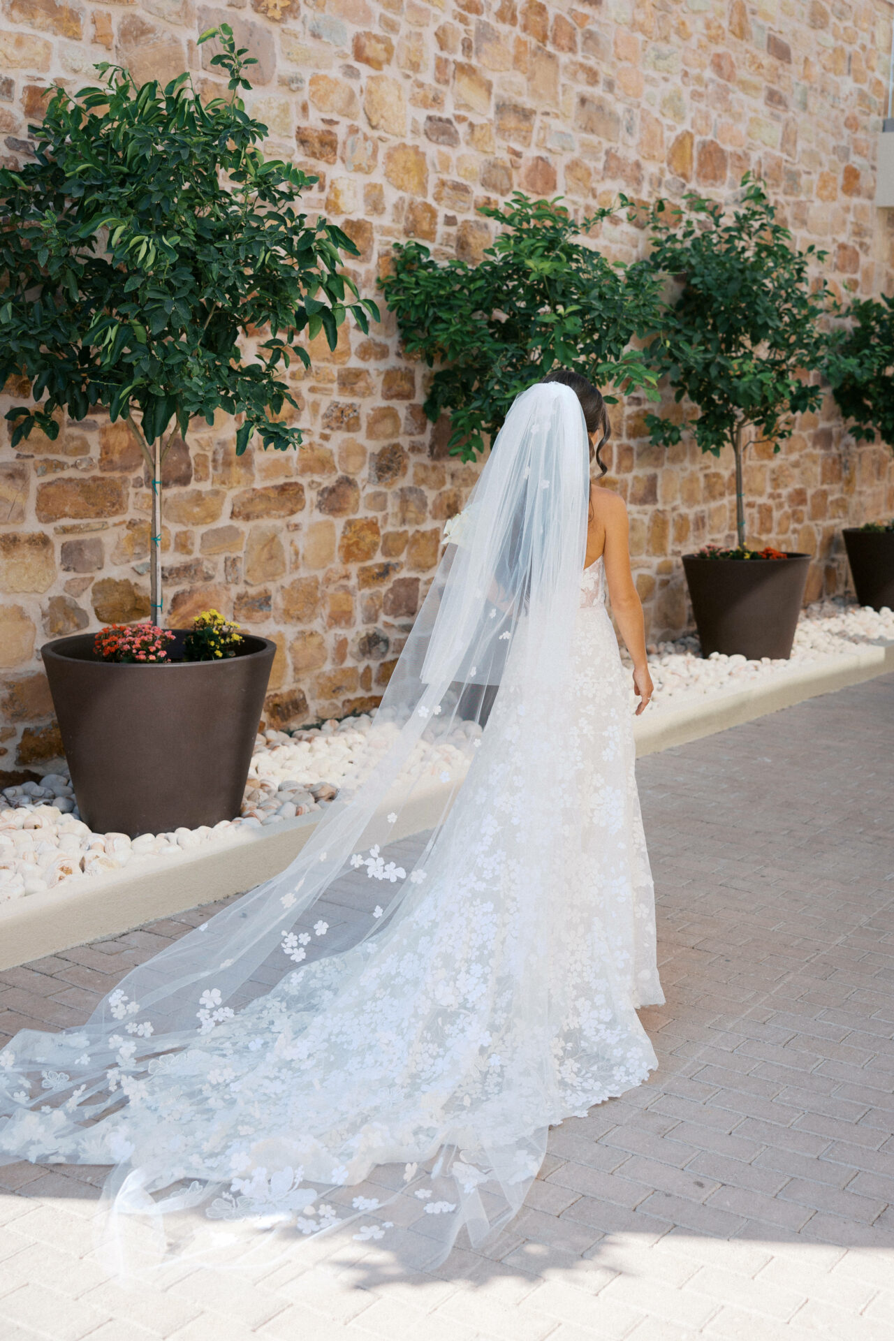 Bride in floral gown walks away by stone wall.