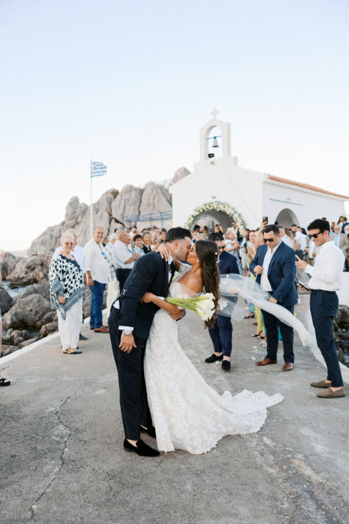 Bride and groom kiss at chapel, Greek flag waves.