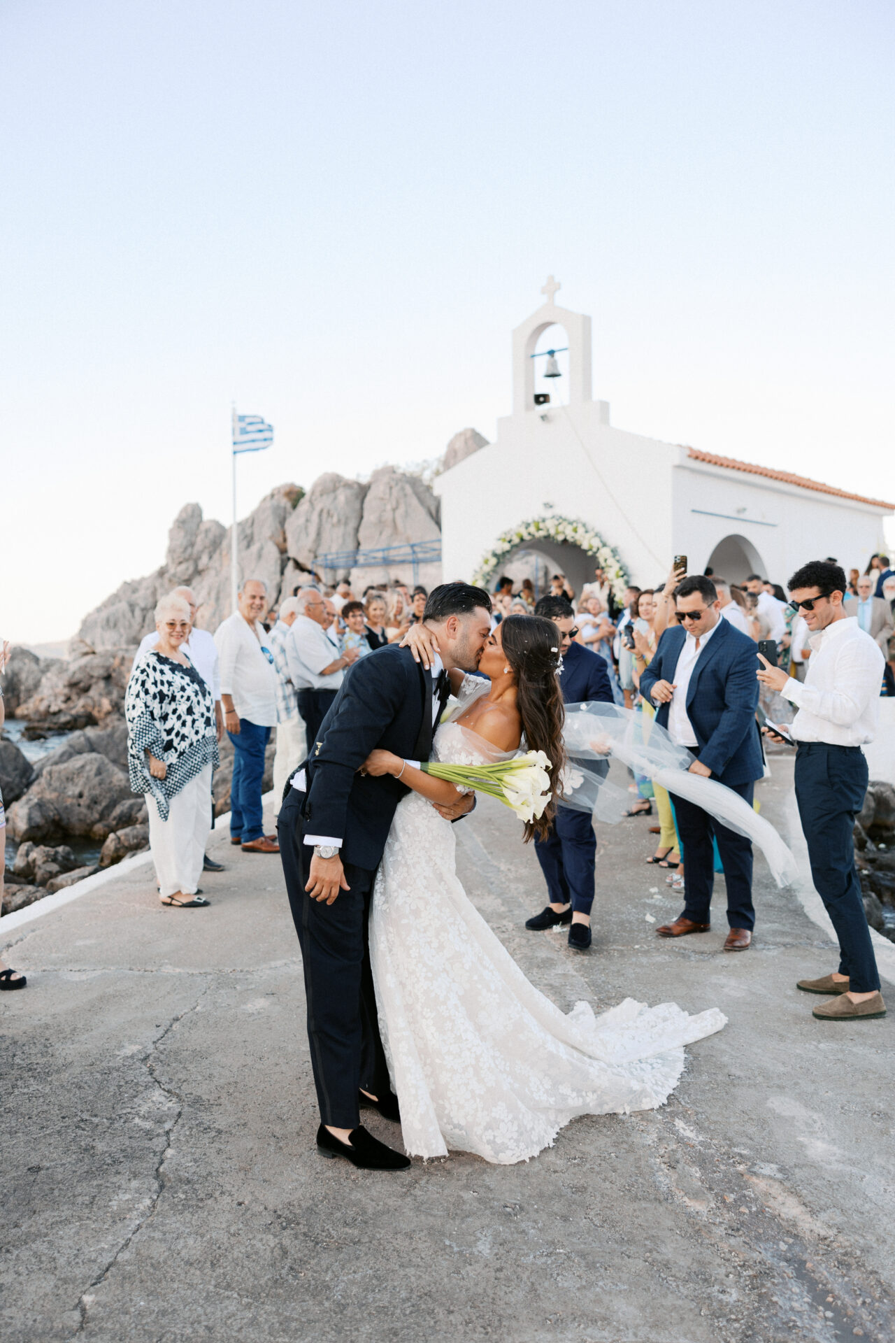 Bride and groom kiss at chapel, Greek flag waves.