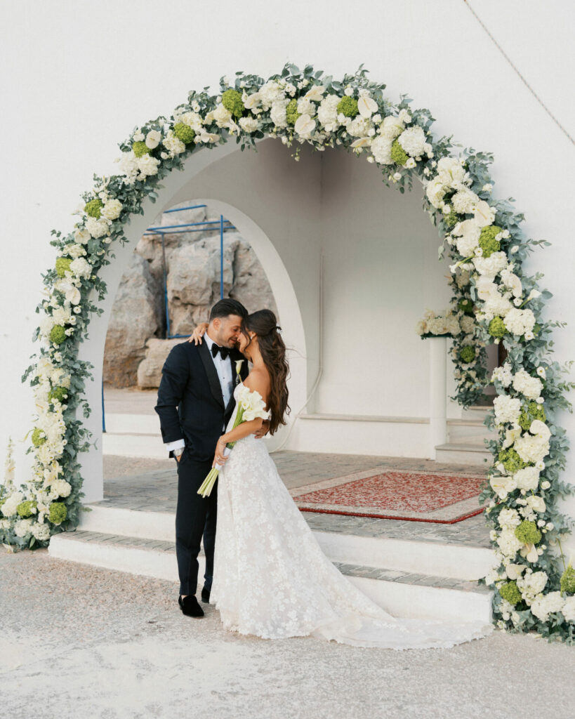 Bride and groom kiss under floral arch outdoors.