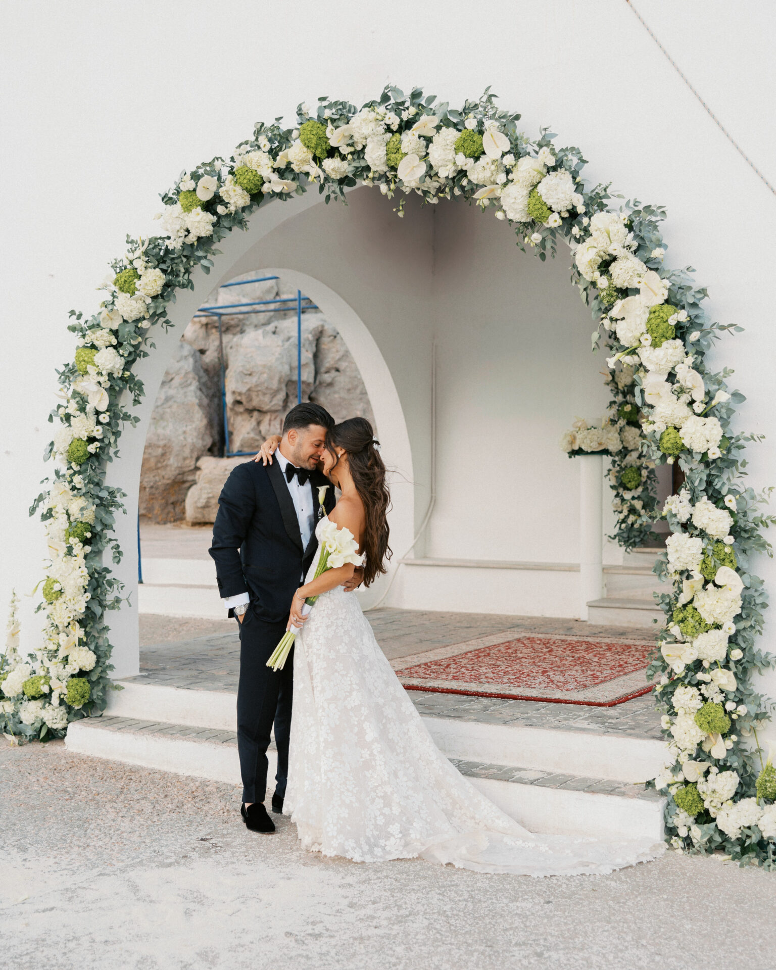 Bride and groom kiss under floral arch outdoors.