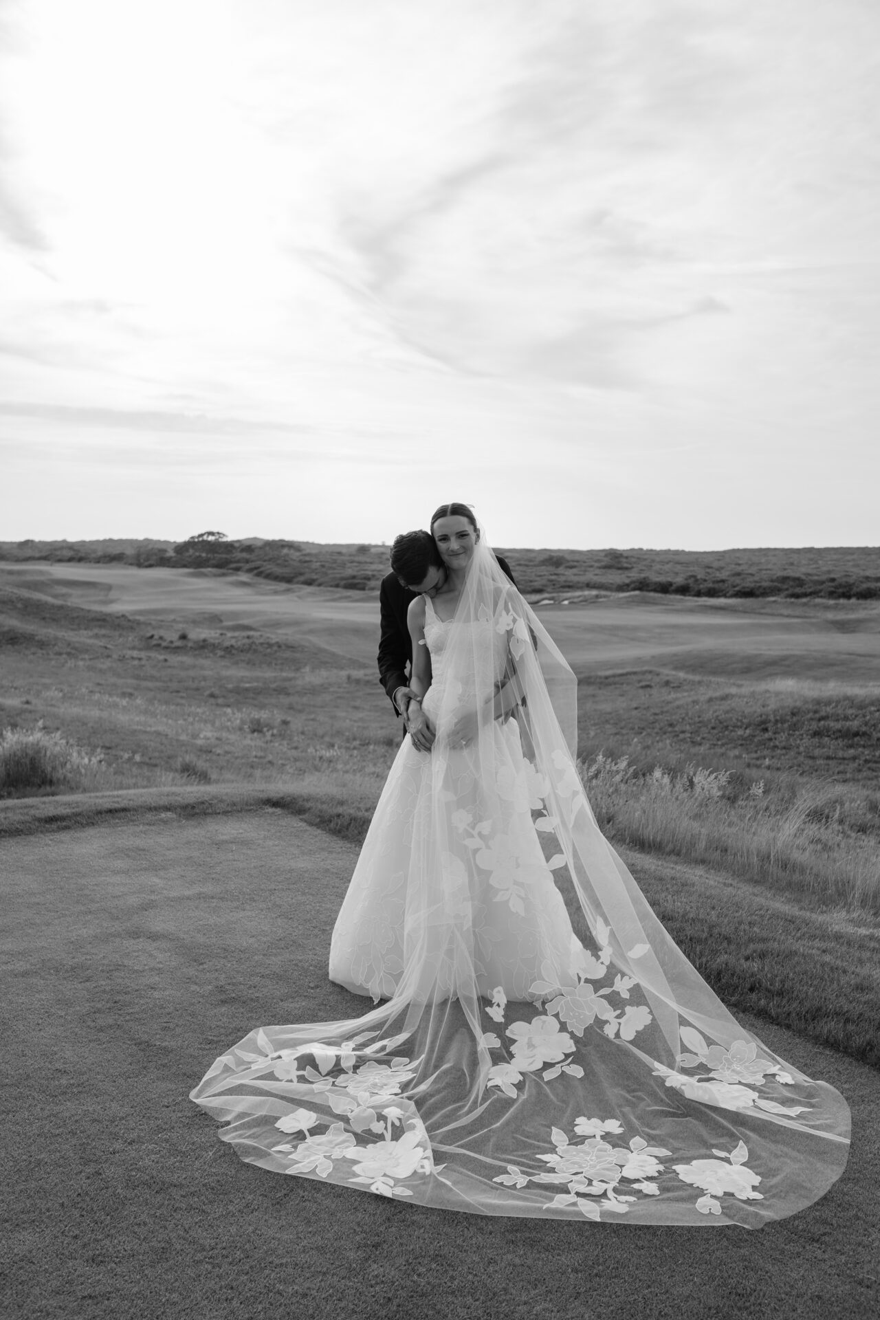 Bride and groom embrace on grassy hillside.